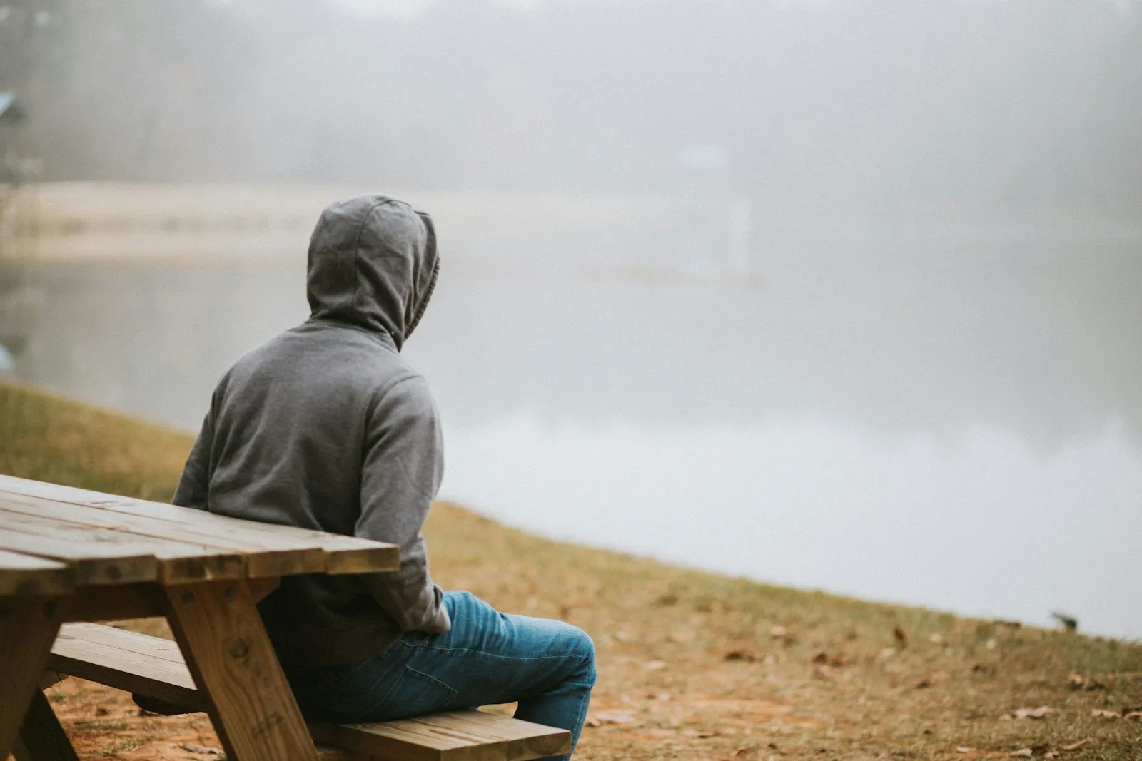 Un adolescent en sweat à capuche, assis seul face à un paysage de brume au bord de l’eau