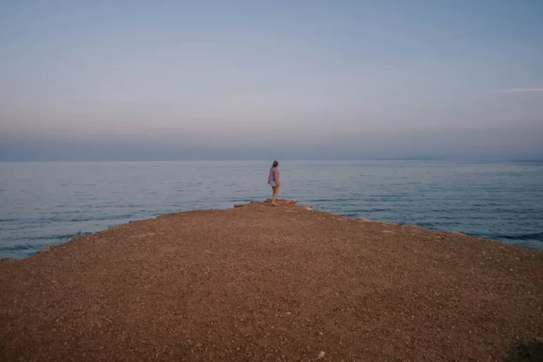 Une femme seule au bout d’une avancée de terre face à la mer, immobile avant de faire un pas