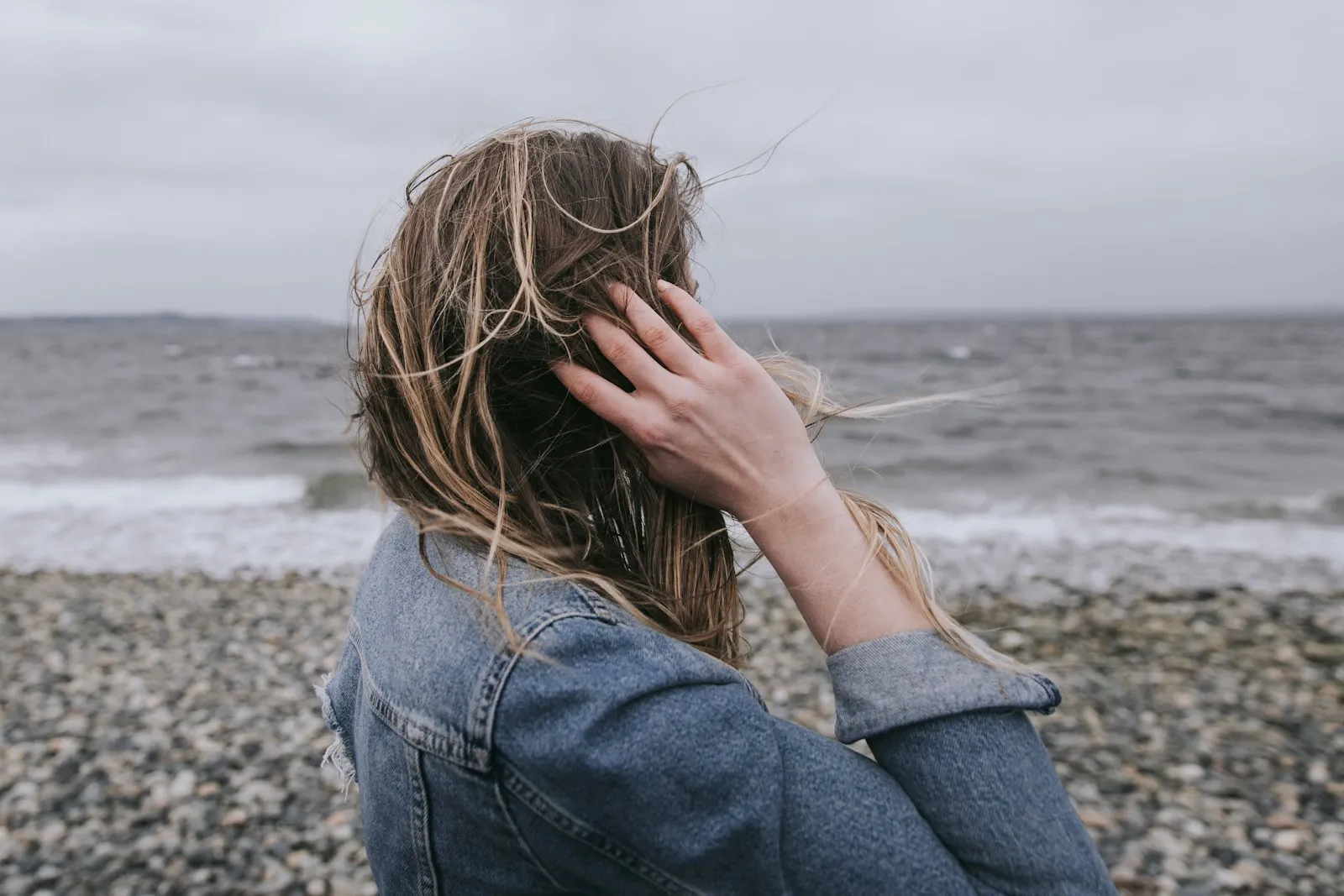 Une femme de dos, cheveux au vent, face à la mer sur une plage de galets